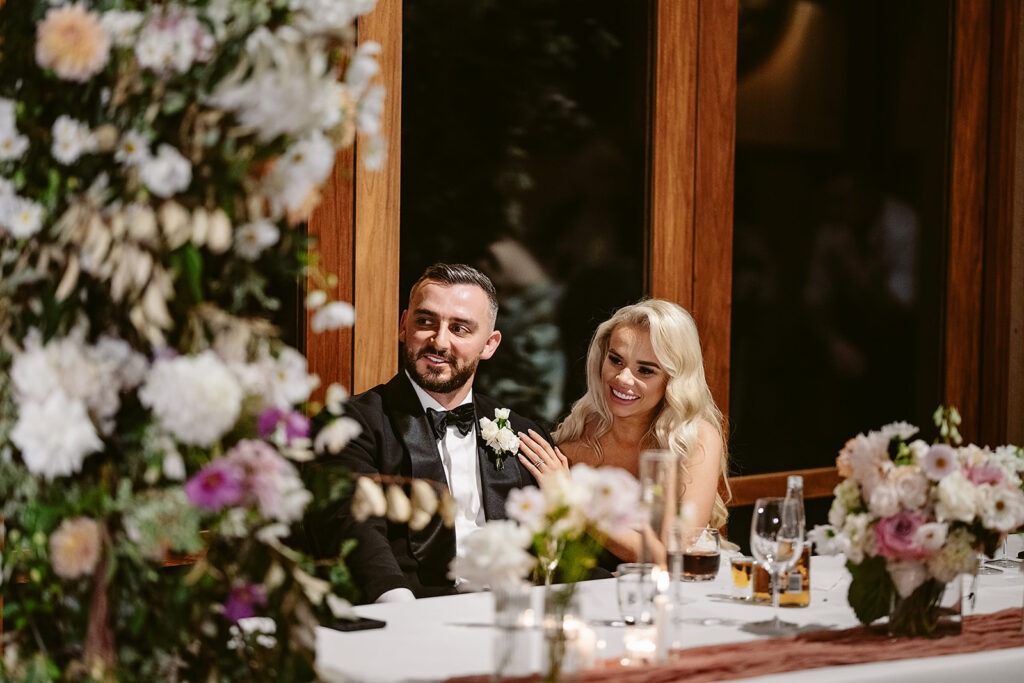 Image of bride & groom at wedding reception, looking towards their guests from the bridal table, happy, lovingly and relaxed. 